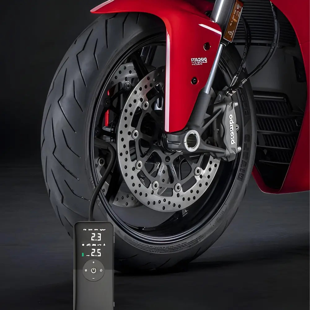 A imagem mostra uma roda dianteira de uma motocicleta vermelha, com um inflador de pneus portátil conectado à válvula do pneu. O visor digital do inflador exibe a pressão de 2.3 e 2.5 psi. A moto e o inflador estão em um fundo escuro, destacando os detalhes do pneu e do inflador.